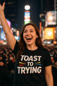 A person with long brown hair is smiling widely and raising one hand in a celebratory manner. They are wearing a black T-shirt with colorful text that reads 