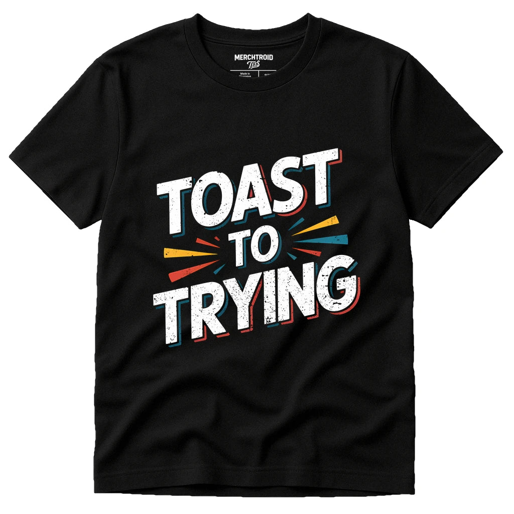 A black T-shirt with the phrase "TOAST TO TRYING" printed in bold white letters, accented with colorful lines in yellow, blue, and red around the text. The shirt label reads "MERCHTROID."
