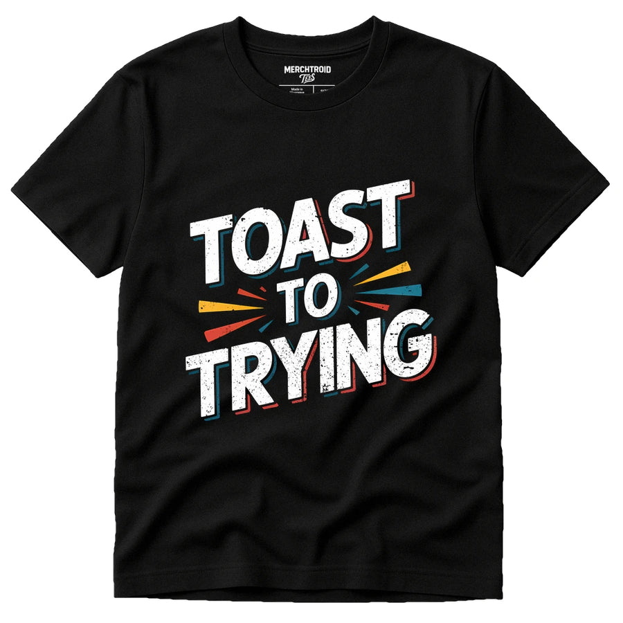 A black T-shirt with the phrase "TOAST TO TRYING" printed in bold white letters, accented with colorful lines in yellow, blue, and red around the text. The shirt label reads "MERCHTROID."