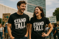 A man and a woman are wearing matching black T-shirts that read “STAND TALL.” They are outdoors, smiling and laughing, with a group of people in the background holding protest signs reading 