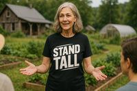 An older woman with gray hair stands outdoors, smiling and gesturing with her hands. She's wearing a black T-shirt with the text 