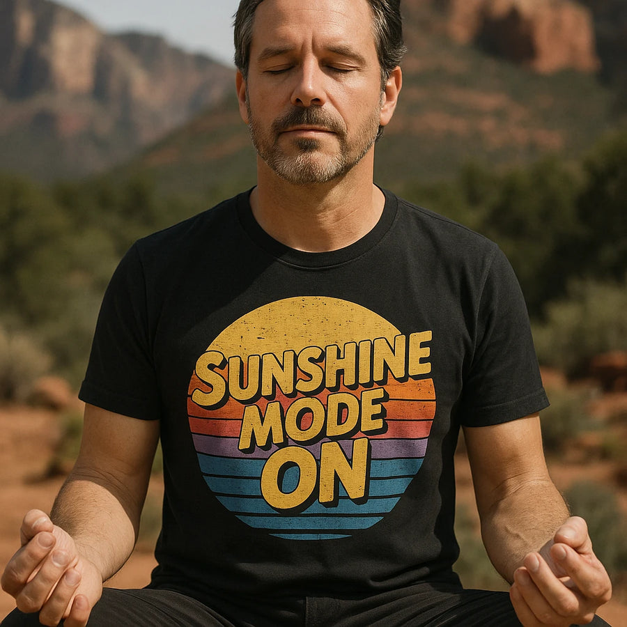 A man meditates outdoors in a serene environment, with his eyes closed and hands resting on his knees. He is wearing a black t-shirt with the text "SUNSHINE MODE ON" over a sunset graphic. The background features a landscape with trees and rocky formations.