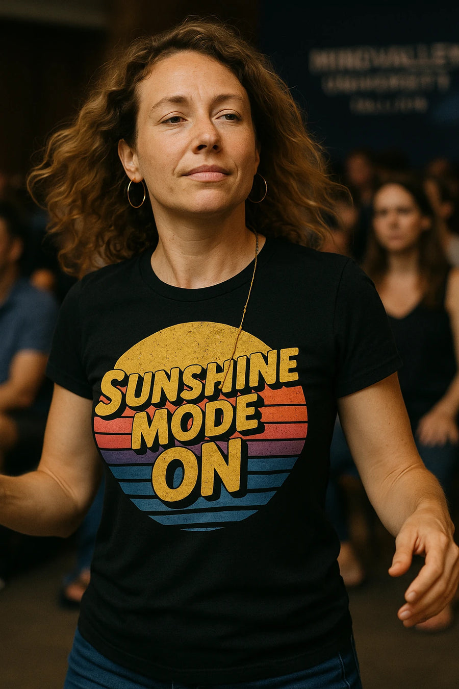 A person with curly brown hair wearing hoop earrings and a black t-shirt with colorful text "SUNSHINE MODE ON." They are in an indoor setting, with blurred people in the background.