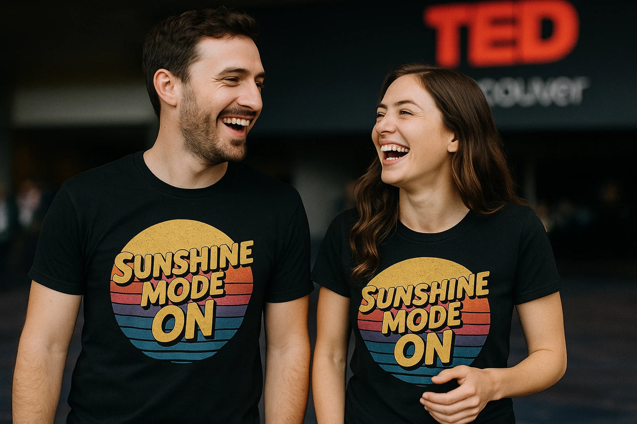 A man and a woman are smiling and laughing together, both wearing black t-shirts with the text "SUNSHINE MODE ON" in bold, colorful letters. They are standing in front of a dark background with blurred text that reads "TED" partially visible.