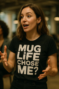 A person with long hair is wearing a black T-shirt with bold white text. They have an expressive pose with hands gesturing outward, and their mouth is open as if speaking. The background is blurred, suggesting an indoor setting.