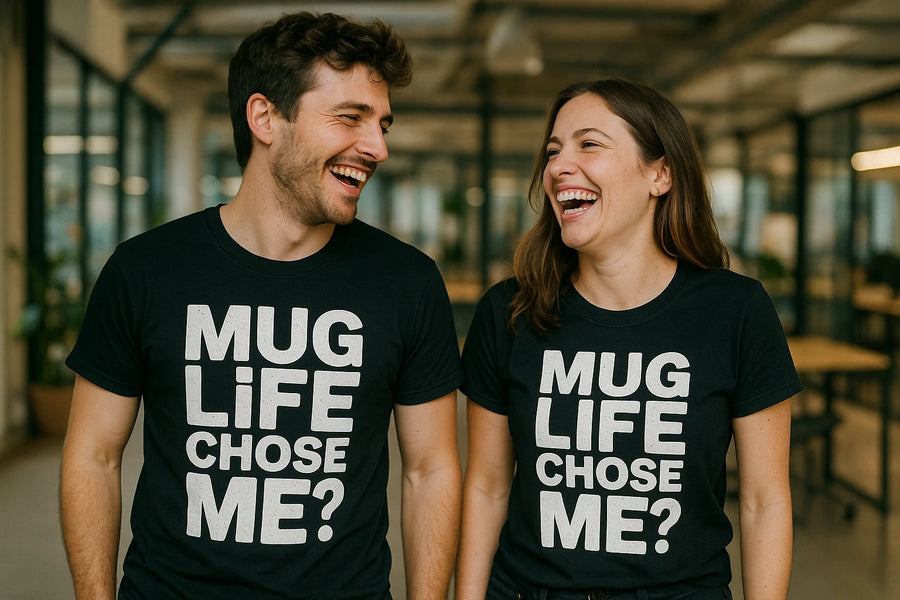 A man and a woman are standing side by side, both laughing and wearing matching black T-shirts with the text "MUG LIFE CHOSE ME?" printed in large white letters. They are in an indoor setting with glass walls and plants in the background.