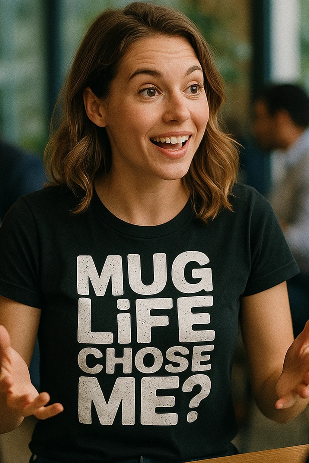 A person with shoulder-length brown hair is smiling and animatedly using their hands. They are wearing a black t-shirt with bold white text. The background is blurred, showing an indoor environment.
