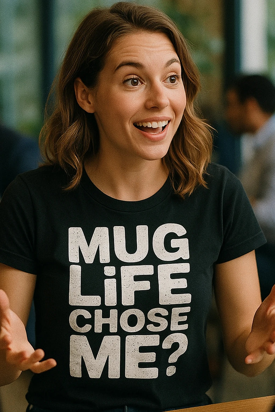 A person with shoulder-length brown hair is smiling and animatedly using their hands. They are wearing a black t-shirt with bold white text. The background is blurred, showing an indoor environment.