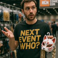 A person with short dark hair and a beard is standing in a convention setting, wearing a black shirt with the text 