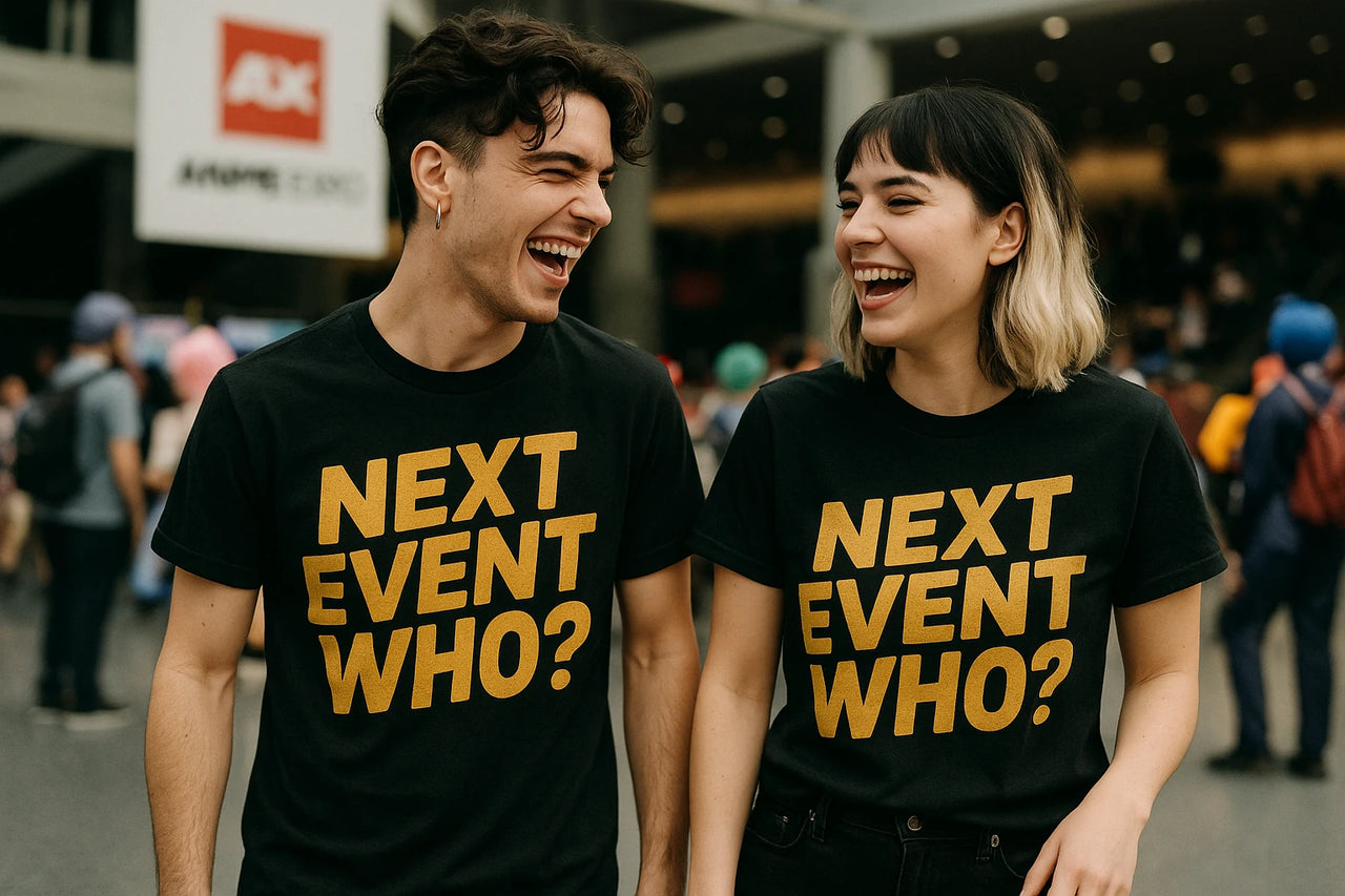 Two people are standing and laughing together. They are wearing matching black T-shirts with the text "NEXT EVENT WHO?" in bold yellow letters. The background is blurred, showing a crowd and an event sign.