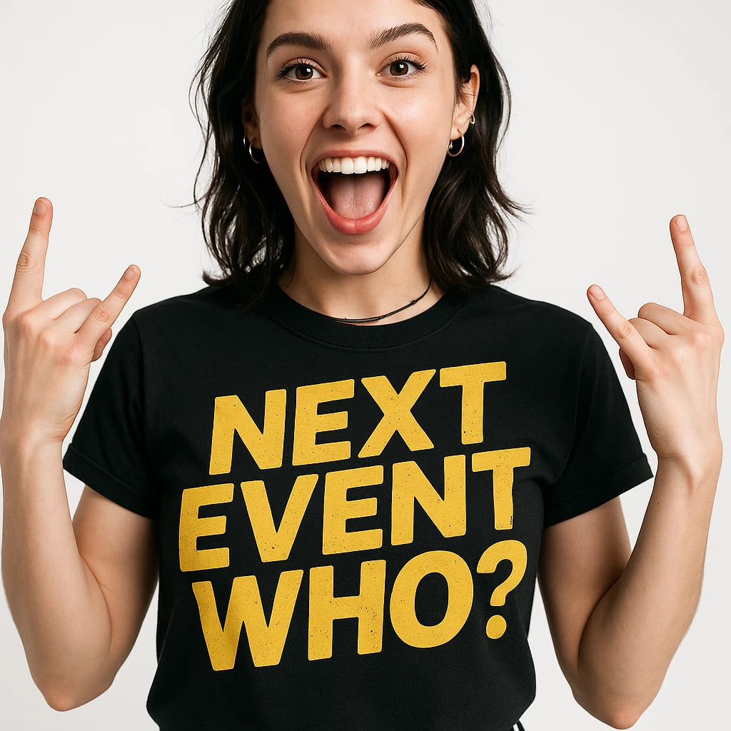 A person with shoulder-length dark hair is wearing a black t-shirt with bold yellow text. They are smiling widely and making a hand gesture often associated with enthusiasm or rock music.