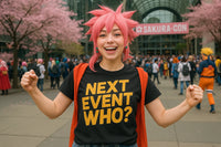 A person with pink spiky hair is smiling and posing energetically at a convention. They are wearing a black T-shirt with the words 