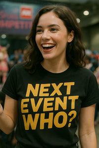 A person with wavy brown hair is smiling widely while wearing a black T-shirt with bold yellow text. The background is blurred, suggesting an indoor event or gathering.