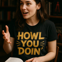 A person with long brown hair is wearing a black T-shirt with yellow text. They appear to be in a library or bookstore setting, holding an open book and gesturing with one hand.