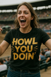 A person with long hair is energetically cheering, wearing a black T-shirt with bold yellow text. They are standing in a stadium with a blurred audience in the background.