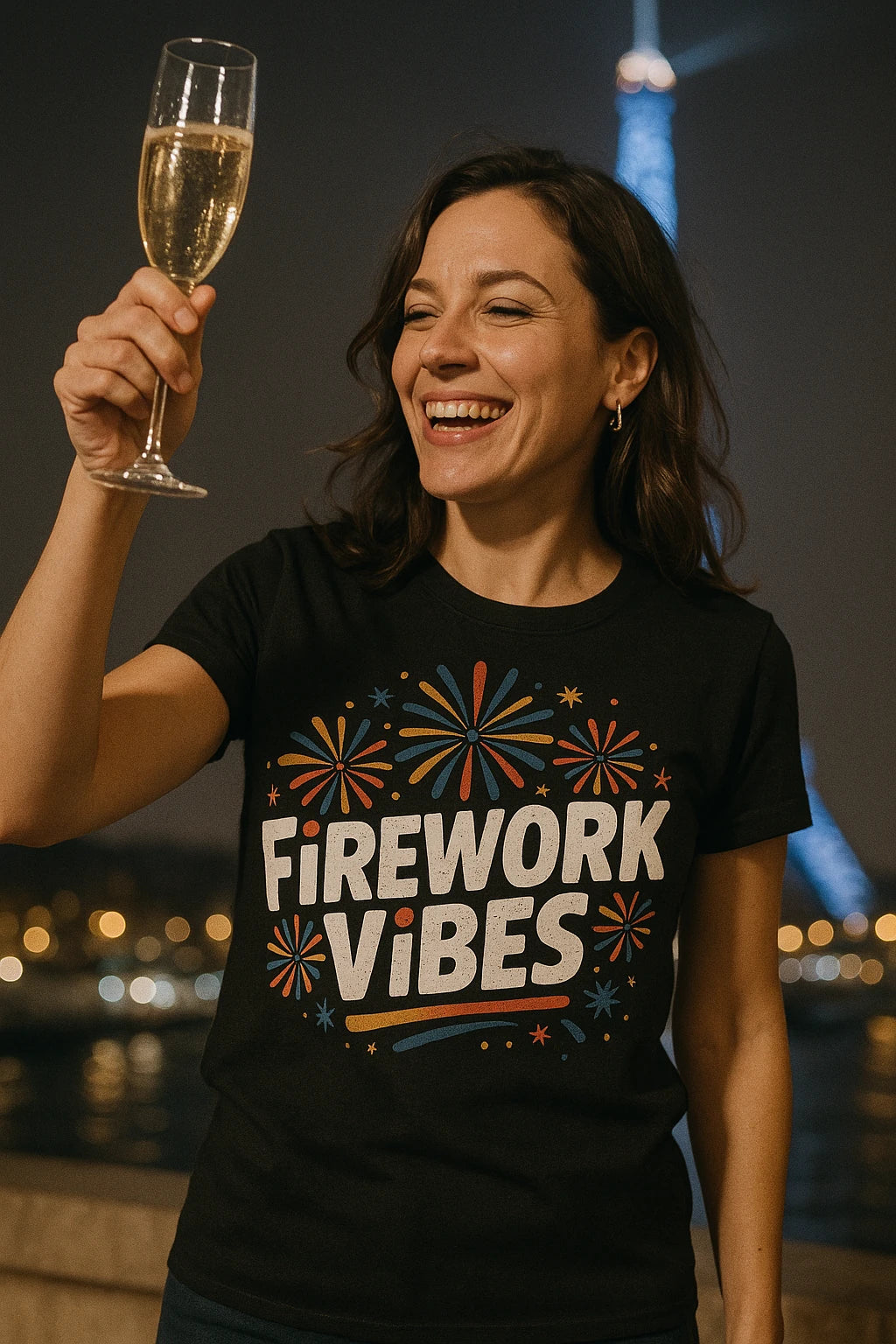 A woman is smiling broadly while holding up a glass of champagne. She is wearing a black t-shirt with colorful firework designs and the text "Firework Vibes." In the background, lights are visible with an iconic tower structure, likely the Eiffel Tower, partially visible. The scene is night-time and appears celebratory.