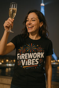 A woman is smiling broadly while holding up a glass of champagne. She is wearing a black t-shirt with colorful firework designs and the text 