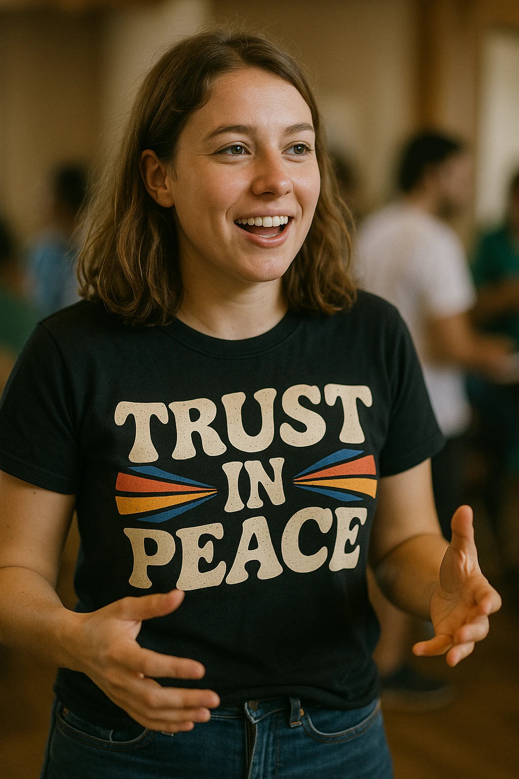 A person with shoulder-length brown hair is smiling and gesturing with their hands. They are wearing a black T-shirt with the text "TRUST IN PEACE" accompanied by colorful graphic elements. The background appears to be a casual indoor setting with people blurred in the distance.