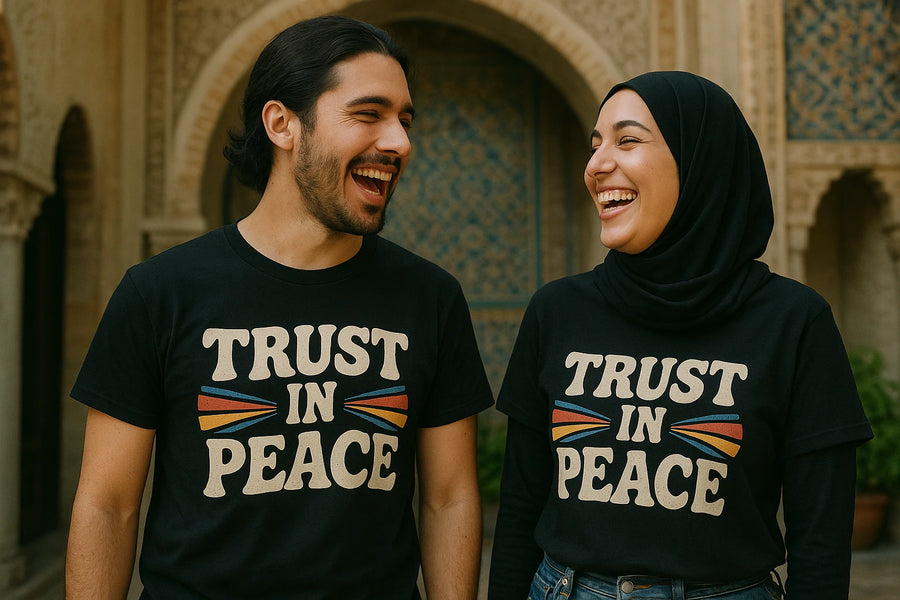 A man with long hair and a beard and a woman wearing a hijab are both laughing and standing in front of an ornate building. They are both wearing black T-shirts with the text "TRUST IN PEACE" and colorful designs.