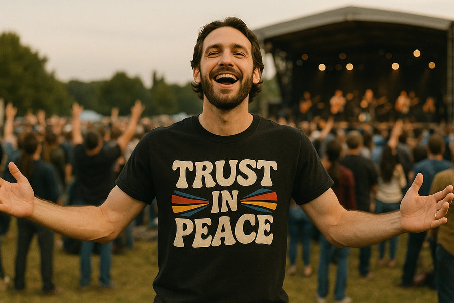 A person with a beard and short hair is smiling broadly and standing outdoors at a music festival. They are wearing a black T-shirt with the text "TRUST IN PEACE" on it, accompanied by colorful graphics. In the background, a crowd and a stage with musicians can be seen.