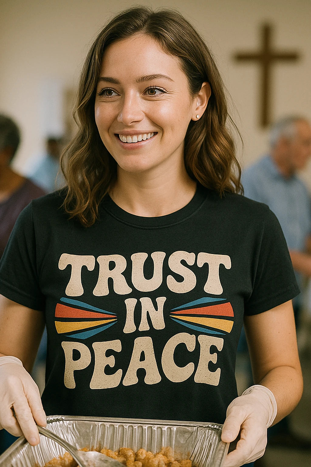 A person with wavy brown hair, wearing a black T-shirt with the text "TRUST IN PEACE" and colorful designs. They are smiling and holding a tray of food while wearing gloves. A blurred cross is visible in the background.