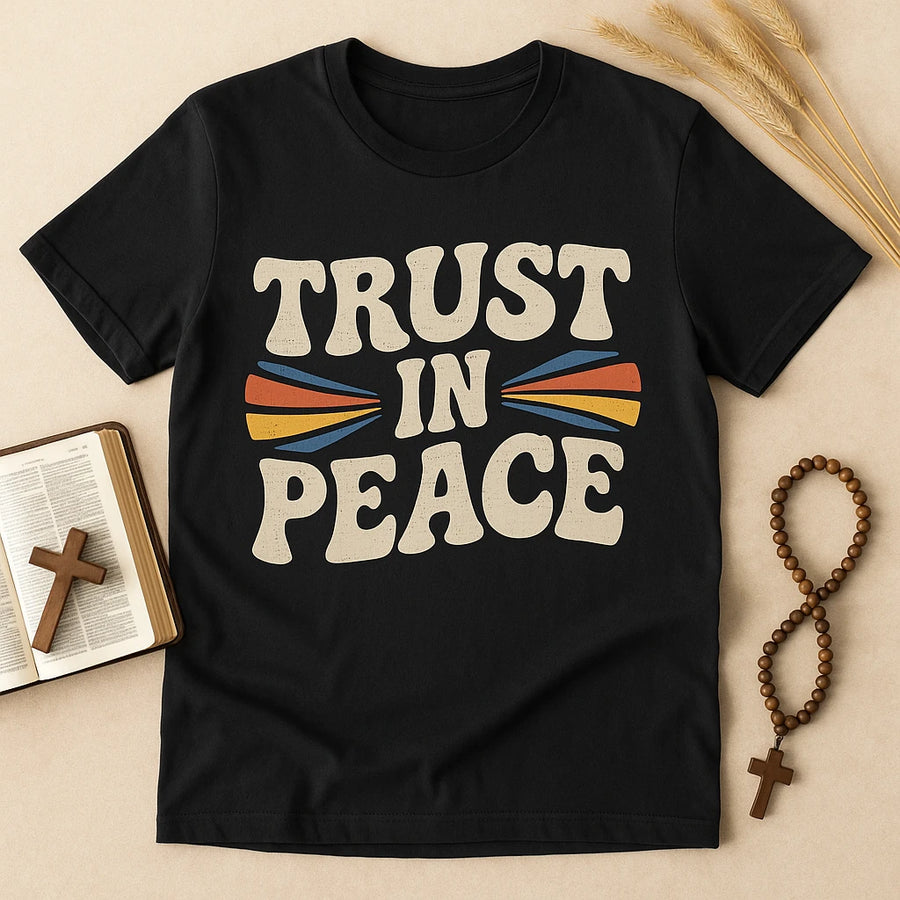 A black T-shirt with large, bold text in a vintage style that reads "TRUST IN PEACE." The design features colorful accents with red, blue, and yellow lines resembling arrows pointing inwards. The shirt is laid out flat on a beige surface, next to a wooden cross on top of an open book, a wooden rosary, and dried wheat stalks.