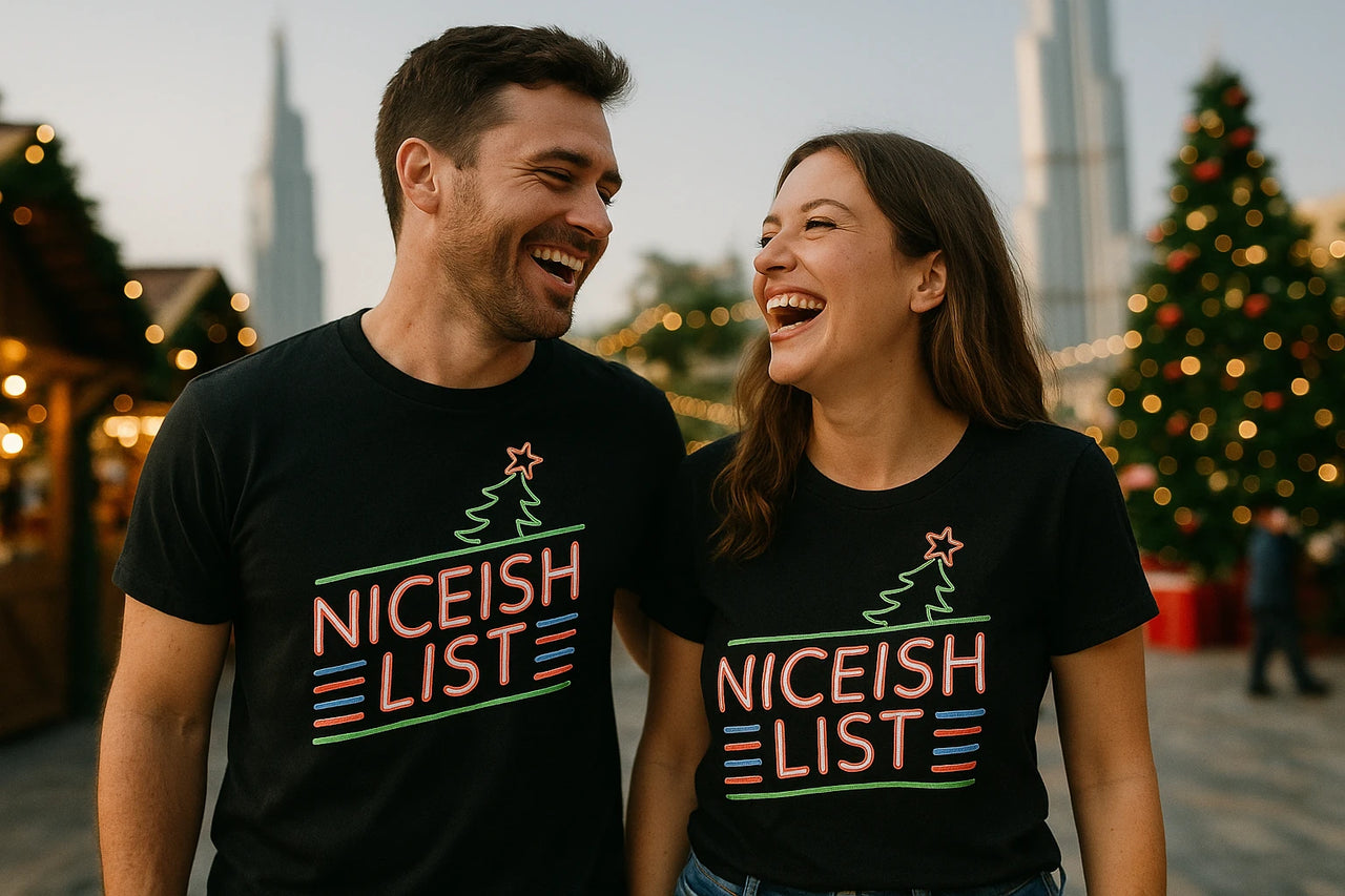A man and a woman are standing close together, laughing while wearing matching black t-shirts with colorful text "NICEISH LIST" and a graphic of a tree with a star on top. They appear to be outdoors, possibly at a festive event, with a decorated Christmas tree and lights in the background.