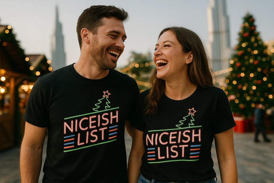 A man and a woman are standing close together, laughing while wearing matching black t-shirts with colorful text "NICEISH LIST" and a graphic of a tree with a star on top. They appear to be outdoors, possibly at a festive event, with a decorated Christmas tree and lights in the background.