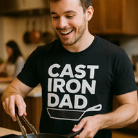 A person with short hair and a beard is smiling while holding a pair of tongs and cooking with a skillet. They are wearing a black t-shirt with the text 