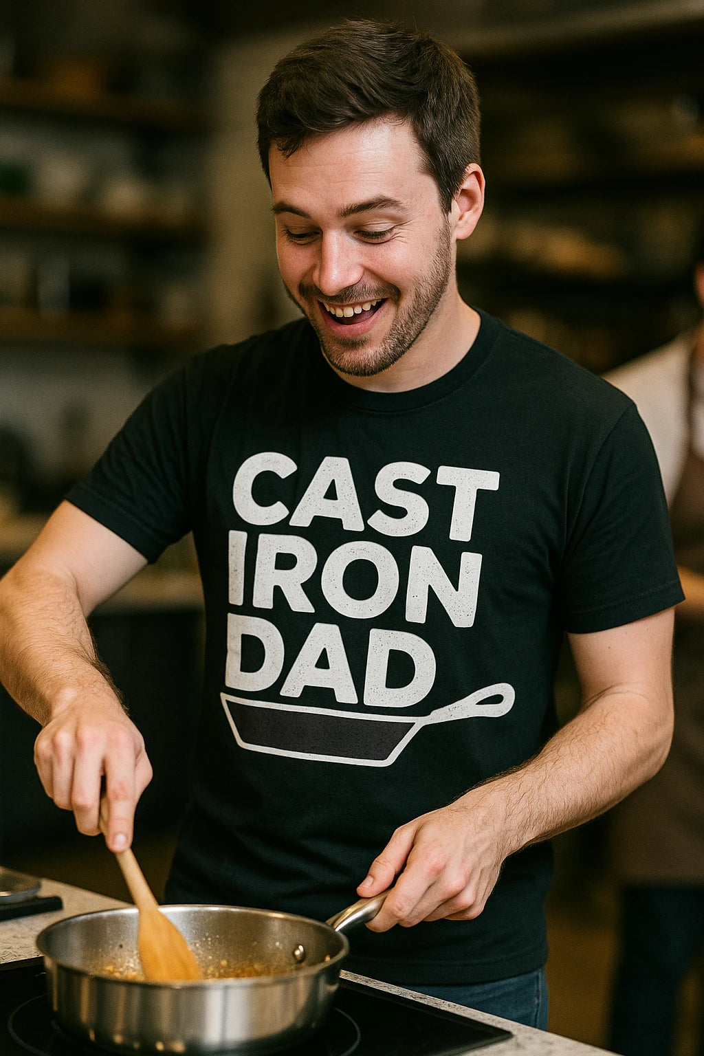 A person with short brown hair and facial stubble is smiling while cooking with a wooden spoon in a kitchen. They are wearing a black T-shirt with the text "CAST IRON DAD" and an illustration of a cast iron skillet.