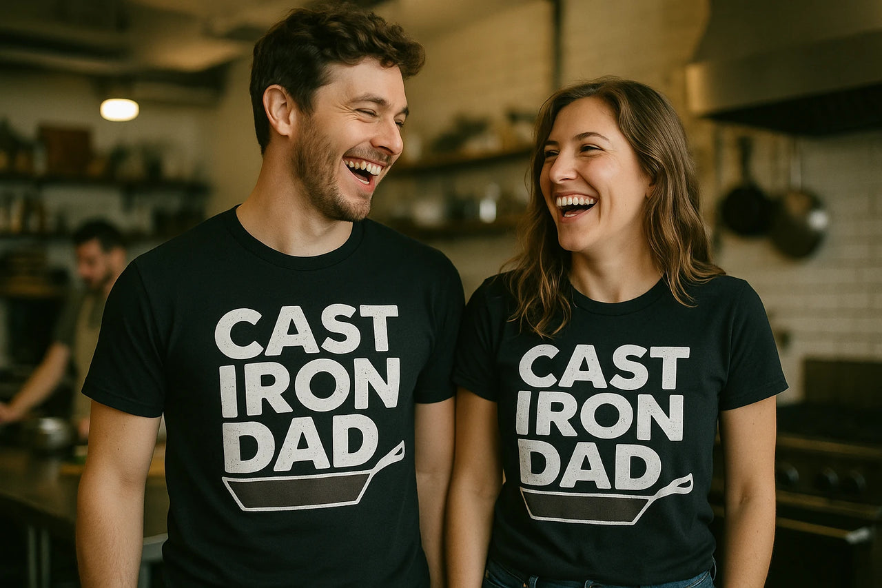 A man and a woman are standing in a kitchen, both wearing matching black t-shirts with the text "CAST IRON DAD" and an illustration of a cast iron skillet. They are smiling and looking at each other, conveying a cheerful and light-hearted mood. The kitchen background is softly blurred, with another person visible in the background.
