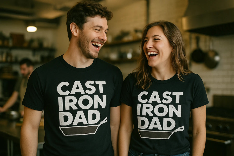 A man and a woman are standing in a kitchen, both wearing matching black t-shirts with the text "CAST IRON DAD" and an illustration of a cast iron skillet. They are smiling and looking at each other, conveying a cheerful and light-hearted mood. The kitchen background is softly blurred, with another person visible in the background.