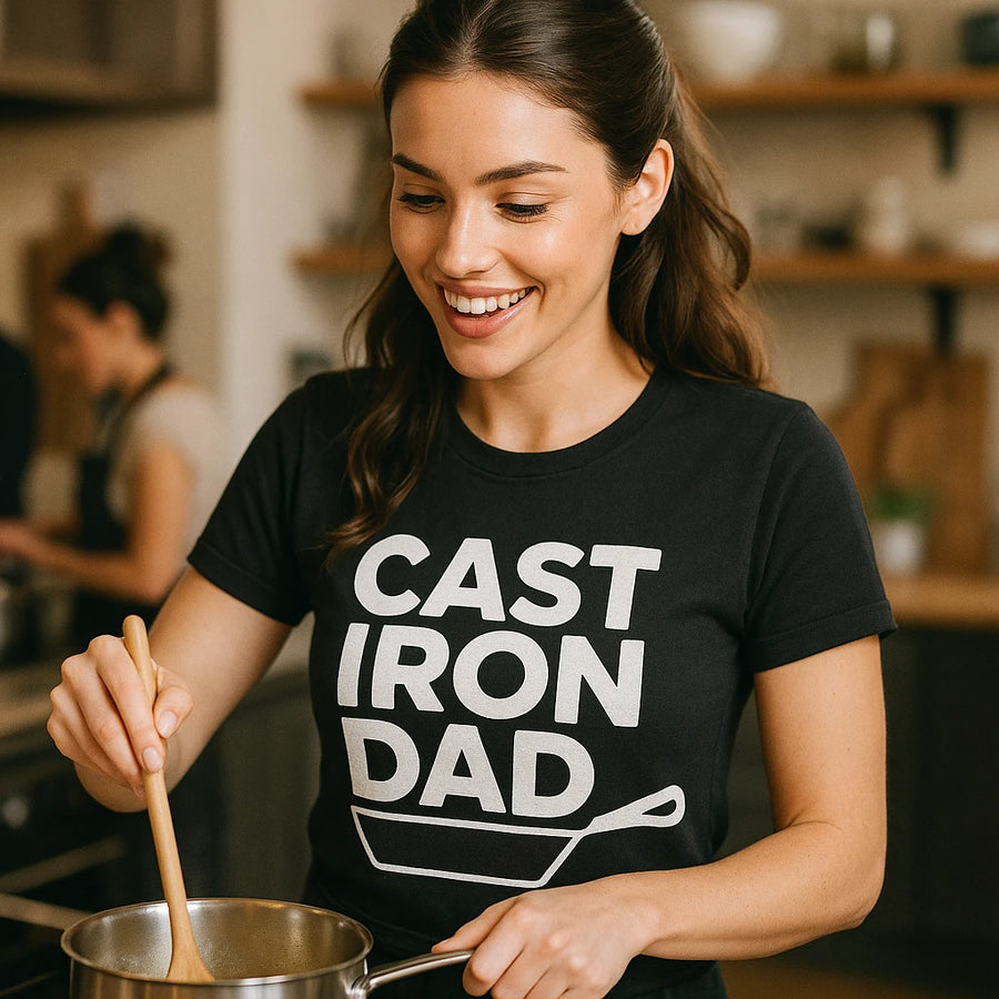 A person with long brown hair is smiling while stirring a pot in a kitchen. They are wearing a black t-shirt with bold white text and a graphic of a frying pan. The background shows a blurred kitchen setting with another person present.
