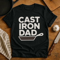 A black t-shirt lying on a wooden surface with a cast iron pan graphic and the words 