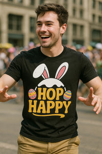 A man is smiling and walking outdoors, wearing a black t-shirt with bunny ears and colorful Easter eggs printed on it along with the text 