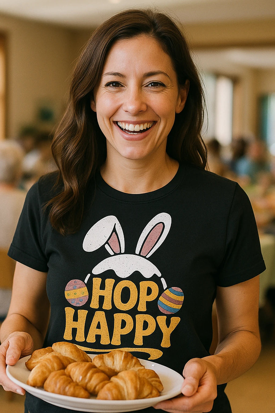 A person with long, dark hair is smiling while holding a plate of croissants. They are wearing a black T-shirt with bunny ears and Easter eggs along with the text "HOP HAPPY." The background is a softly blurred indoor setting, suggesting a social gathering.