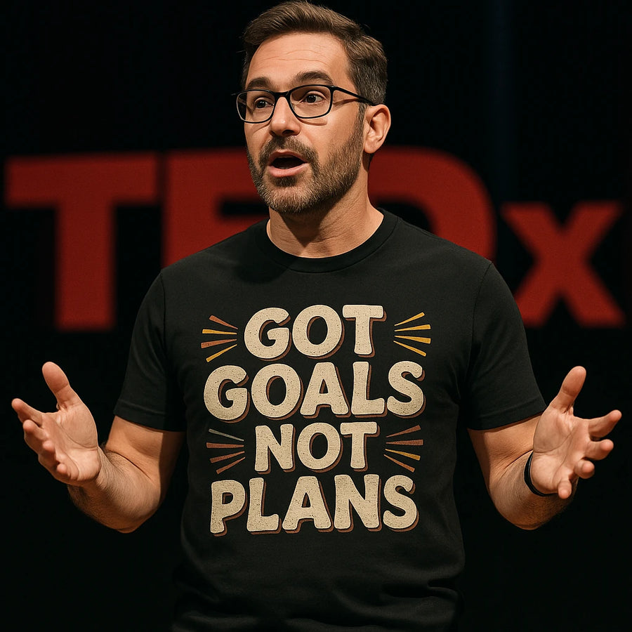 A person wearing glasses and a black T-shirt with the text "GOT GOALS NOT PLANS" is speaking on stage. The background suggests a TEDx event. The individual appears engaged and is gesturing with both hands.