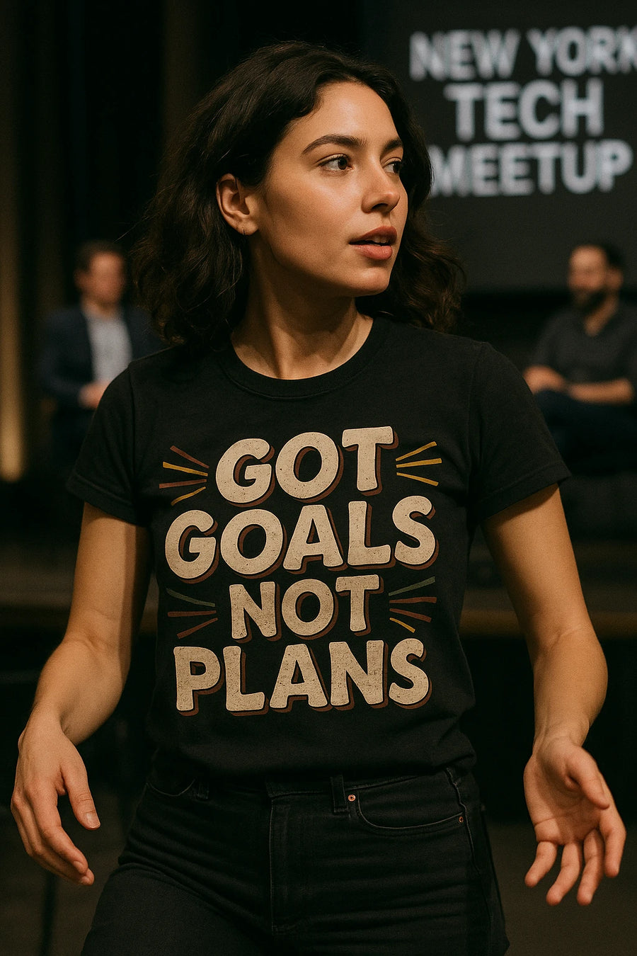 A person with wavy, shoulder-length hair wearing a black t-shirt with text. In the background, there's a blurred setting with a sign reading "NEW YORK TECH MEETUP" and some seated people.