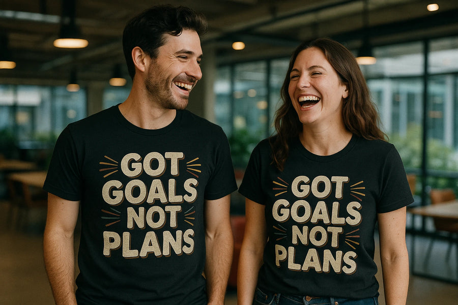 Two people wearing matching black t-shirts with the text "GOT GOALS NOT PLANS" printed on them. They are standing indoors, smiling and laughing, in a well-lit room with tables and windows in the background.
