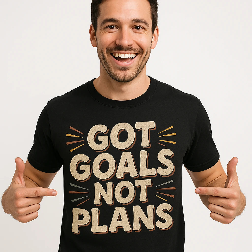 A person smiling broadly and pointing at their black t-shirt with bold text that reads "GOT GOALS NOT PLANS." The background is a plain white, adding emphasis to the t-shirt's message.