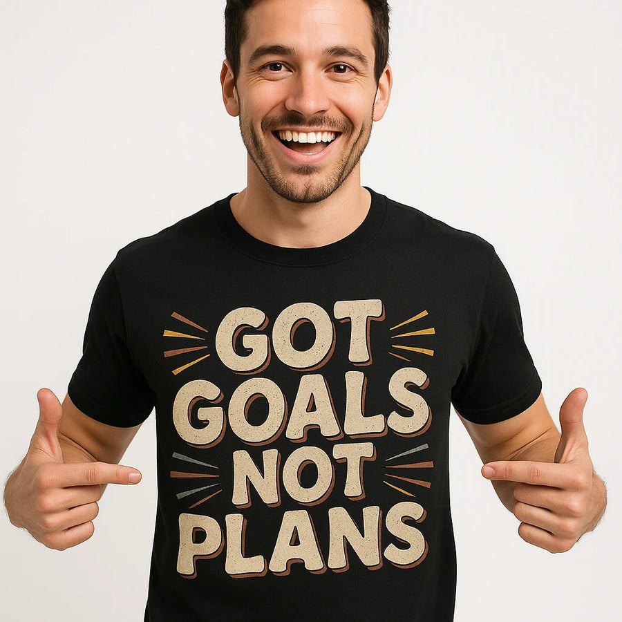 A person smiling broadly and pointing at their black t-shirt with bold text that reads "GOT GOALS NOT PLANS." The background is a plain white, adding emphasis to the t-shirt's message.