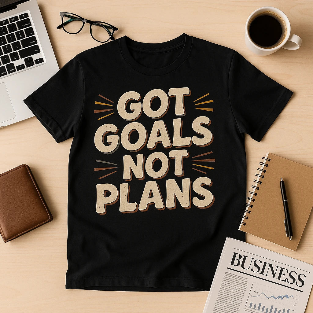 A black t-shirt with the text "GOT GOALS NOT PLANS" displayed on a wooden desk surface. Surrounding the shirt are a laptop, a pair of eyeglasses, a brown leather wallet, a cup of coffee, a notebook with a pen, and a newspaper section labeled "BUSINESS."