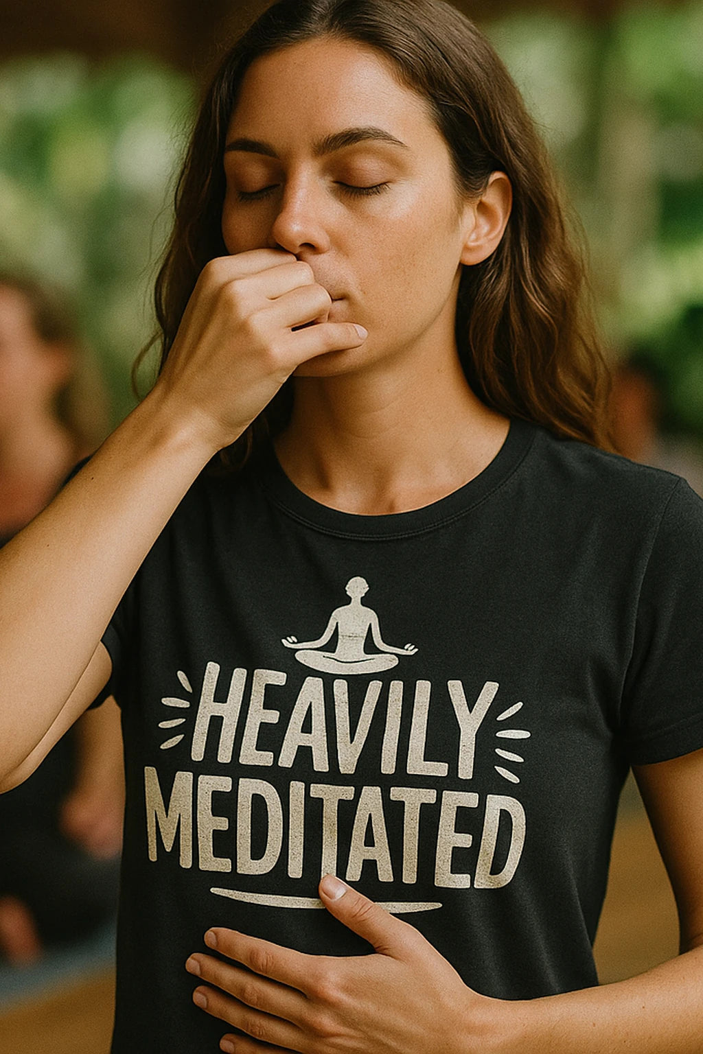 A person with long hair is practicing breathing exercises with one hand on their face and the other on their stomach. They are wearing a black shirt that features a graphic of a meditating figure and the text "HEAVILY MEDITATED." The background is softly blurred, suggesting an indoor setting with natural light.