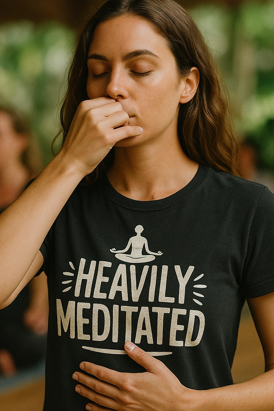 A person with long hair is practicing breathing exercises with one hand on their face and the other on their stomach. They are wearing a black shirt that features a graphic of a meditating figure and the text "HEAVILY MEDITATED." The background is softly blurred, suggesting an indoor setting with natural light.