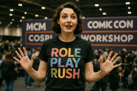 A person with short, wavy hair is standing indoors with people in the background. They are wearing a black T-shirt with the text 