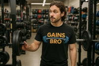 A person with shoulder-length brown hair and a beard is standing in a gym, holding a weight plate on a rack. They are wearing a black T-shirt with the text 