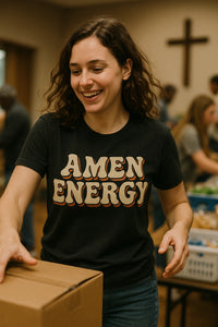A person with curly brown hair is smiling and holding a cardboard box. They are wearing a black t-shirt with the words 