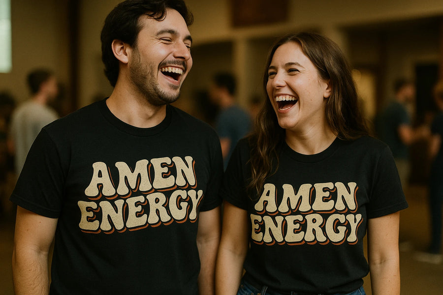A man and a woman are standing next to each other, laughing joyfully. Both are wearing black T-shirts with the phrase "AMEN ENERGY" printed in bold letters. The background is a blurred indoor setting, possibly at a social gathering.