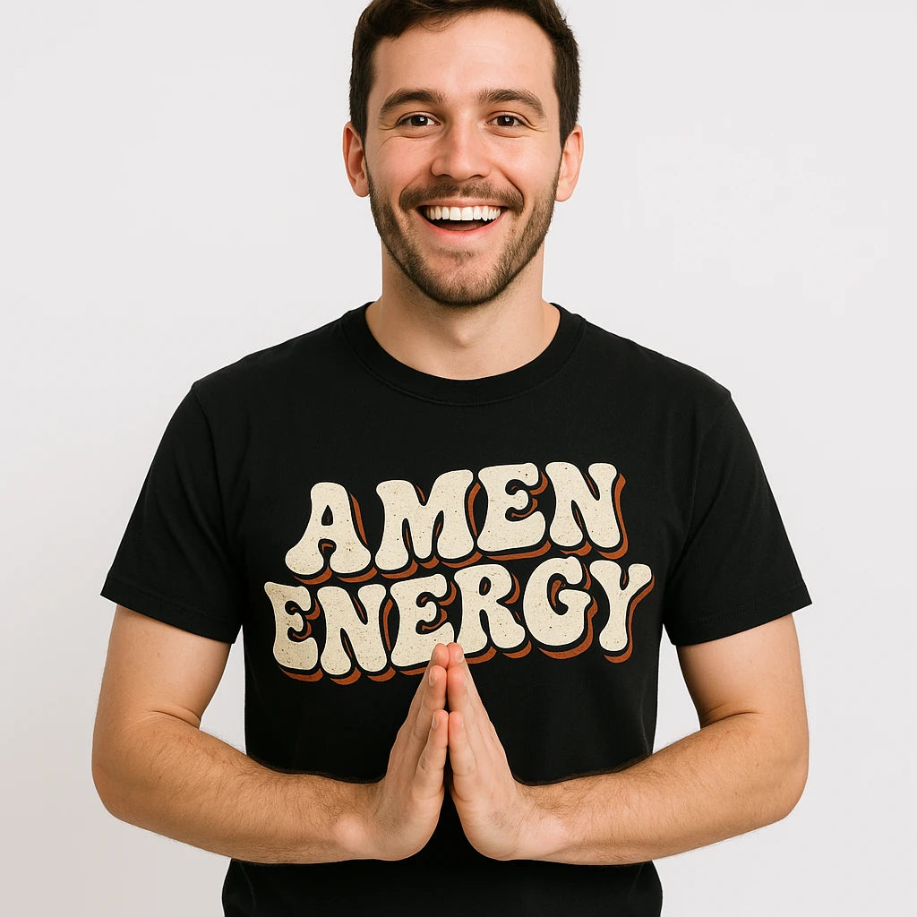 A person with short dark hair and a beard is smiling and standing against a plain background. They are wearing a black T-shirt with the words "AMEN ENERGY" written in bold, stylized letters. The person is holding their hands together as if in a prayer gesture.