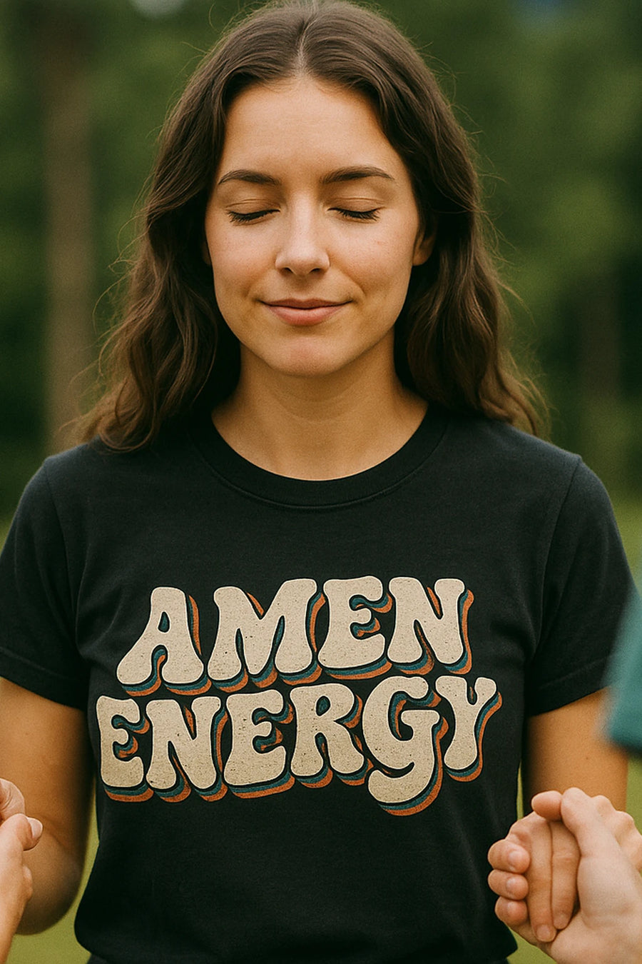 A person with long brown hair is standing outdoors with their eyes closed and a gentle smile, wearing a black shirt with the text "AMEN ENERGY." Their hands are being held by others, suggesting a moment of connection or meditation in a natural setting.